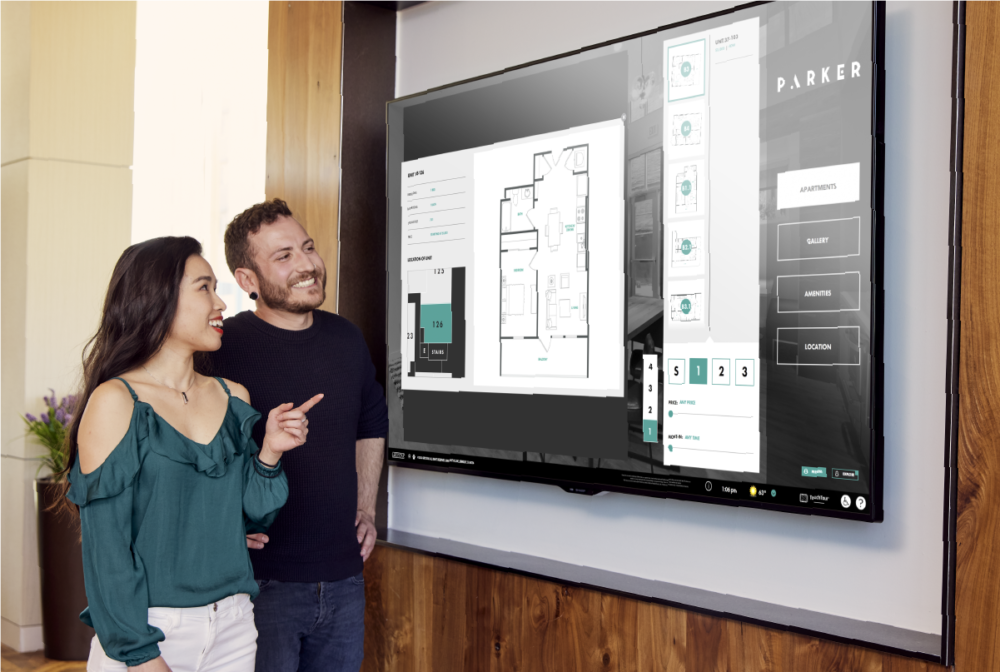Two people smiling while interacting with large wall mounted Touch Tour screen showing software interface with floorplan details.